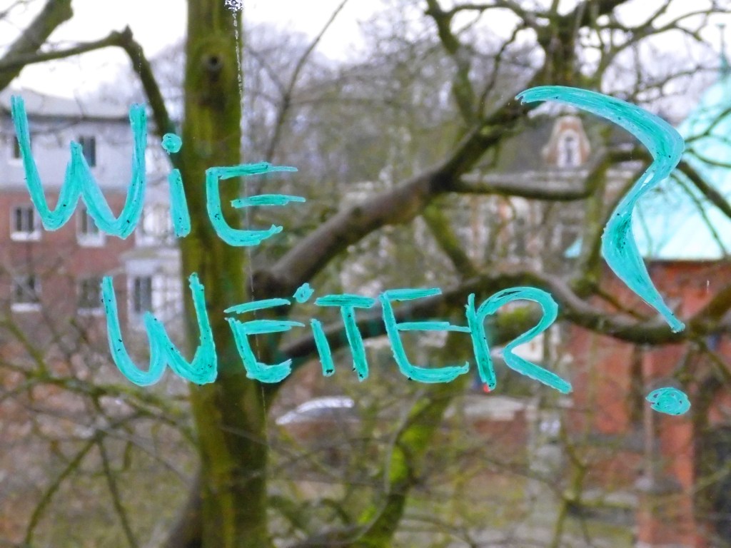 Ein türkisfarbener Schriftzug auf einer Glasscheibe fragt "Wie weiter?" und im Hintergrund ein Baum.