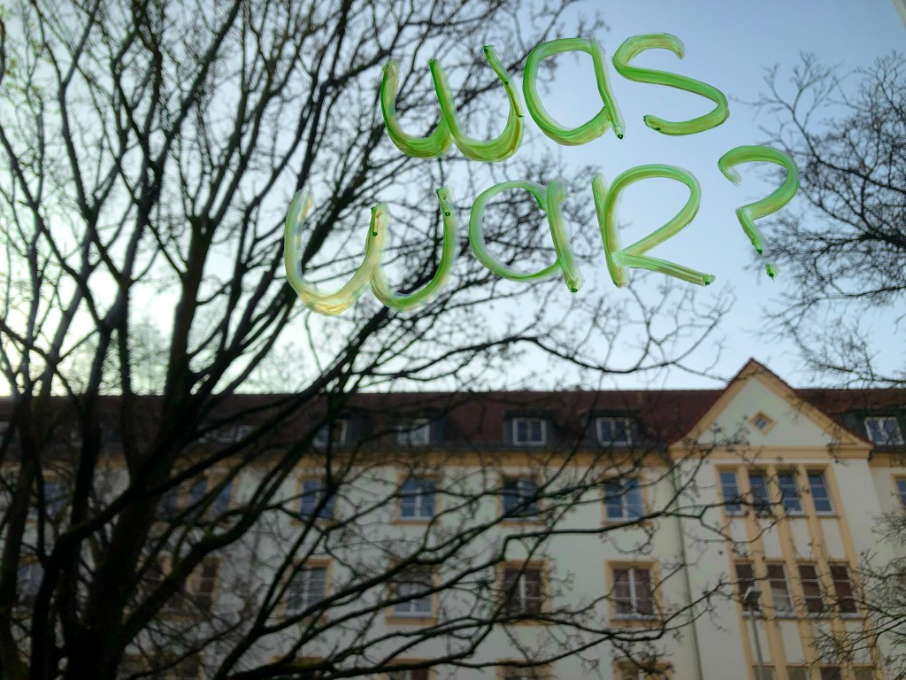 Ein neongrüner Schriftzug an eine Fensterscheibe geschrieben fragt "Was war?", dahinter ein Baum und ein gelbes Haus vor blauem Himmel.