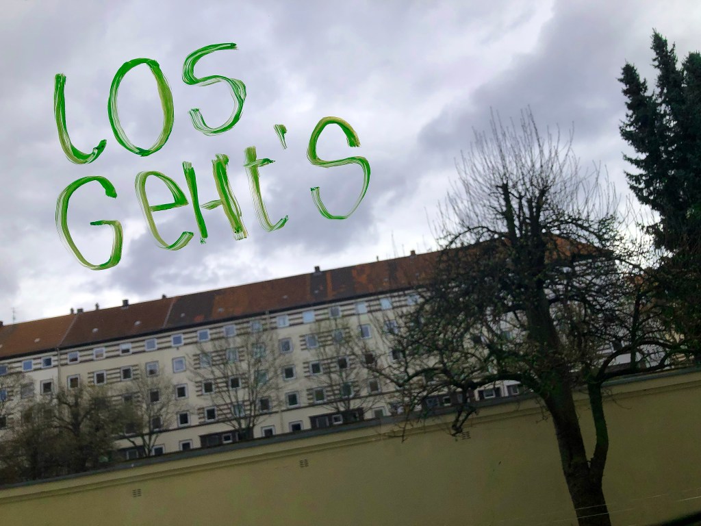 Ein neon-grüner Schriftzug "Los geht's" vor einem wolkigen Himmel, darunter eine Häuserfront und Bäume.