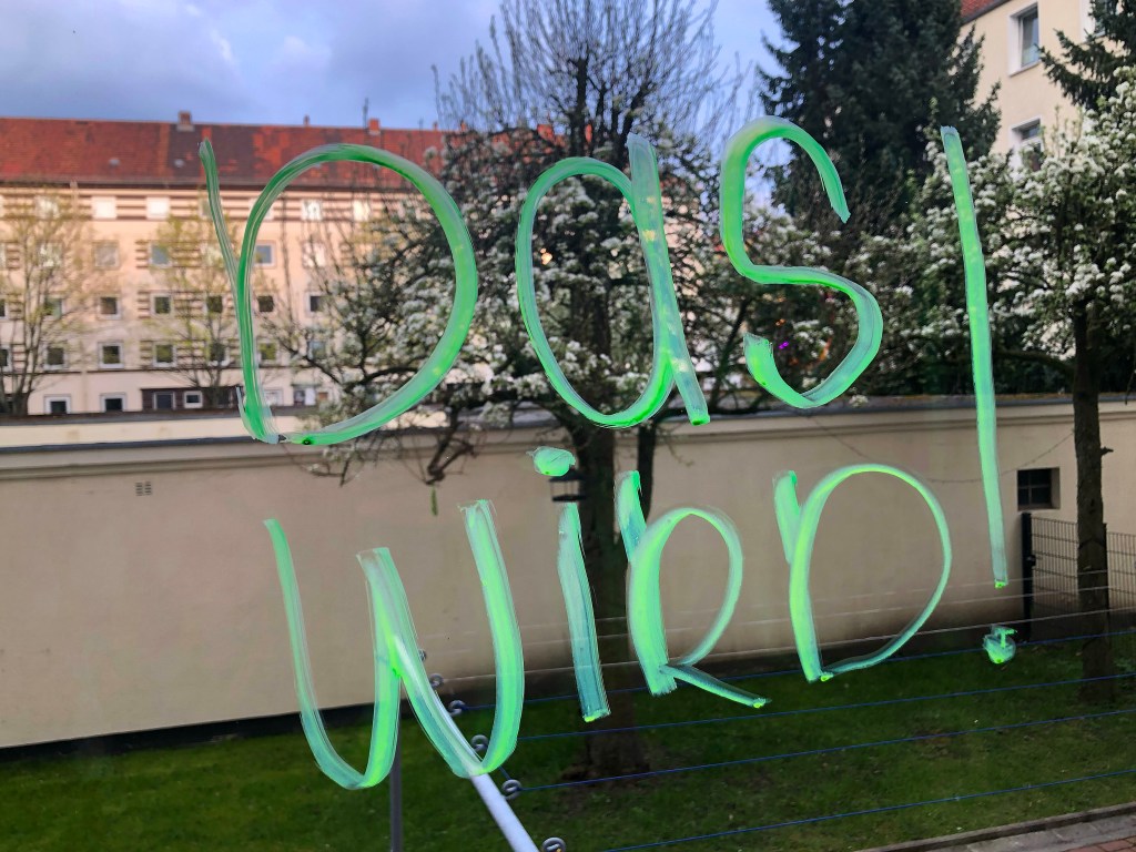 Ein neongrüner Schriftzug "Das wird!" an einer Fensterscheibe, im Hintergrund blühende Kirschbäume und ein Mehrfamilienhaus