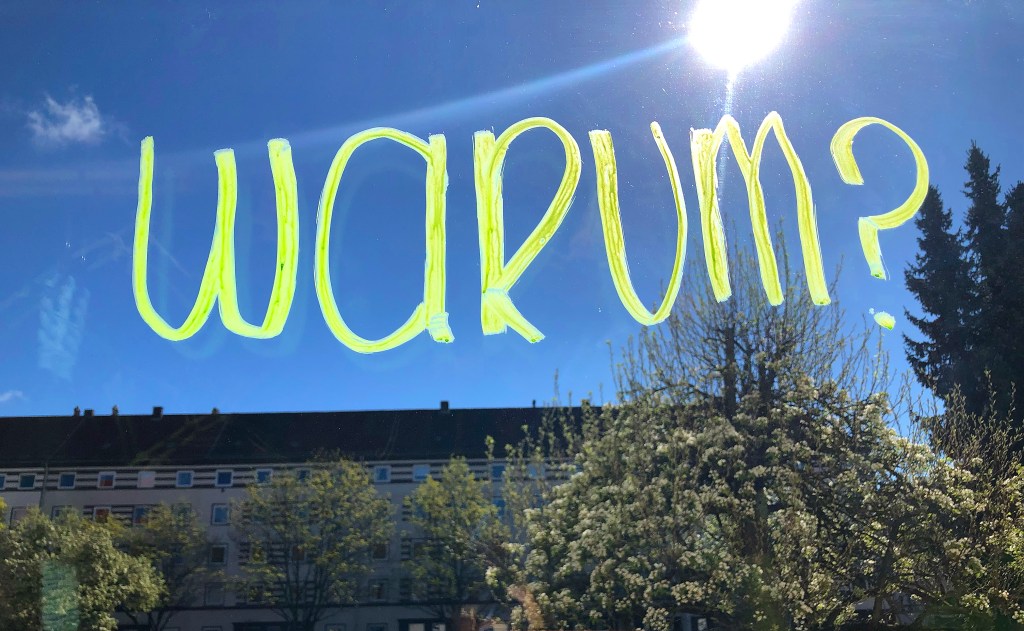 Ein grün-gelber Schriftzug "Warum?" vor einem blauen Himmel mit Sonne, im Hintergrund ein Mehrfamilienhaus und blühende Bäume.