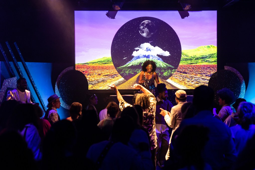 Eine Menschenmenge (von hinten zu sehen) schaut auf eine schwarze Frau auf einer Bühne, im Hintergrund eine Leinwand, die eine grafische Collage mit Weltkugel und einer Landschaft zeigt