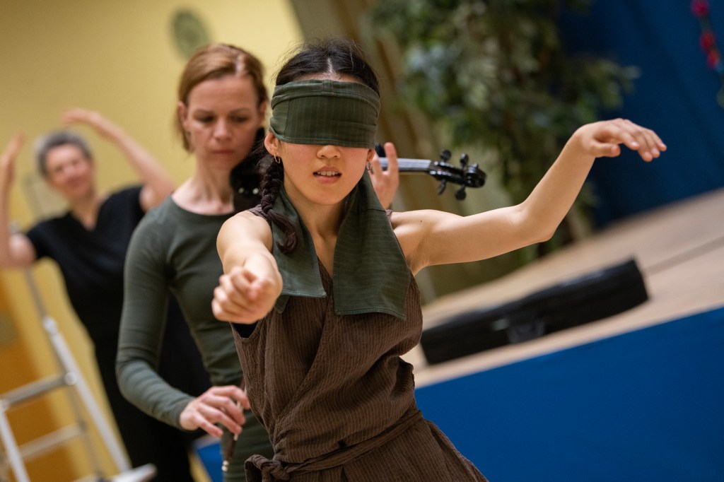 Eine Person im braunen Kleid mit grüner Augenbinde, dahinter steht eine Geigerin, im Hintergrund eine DGS-Dolmetscherin