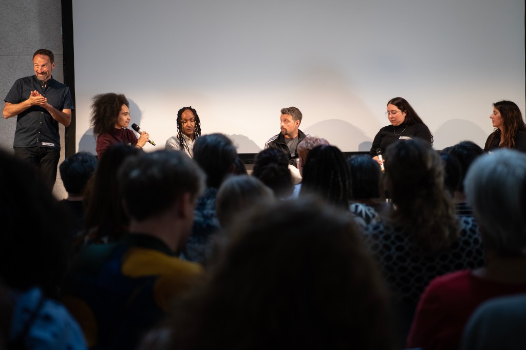 Eine Menschenmenge von hinten schaut auf fünf Menschen, die sich auf einer Bühne sitzend unterhalten, daneben ein DGS-Dolmetscher