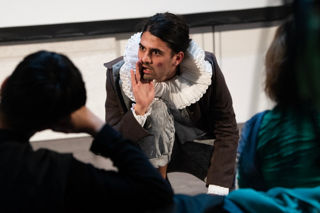 Eine Person of Colour mit rotem Punkt auf der Wange, shakespearescher Halskrause und braunem Jackett