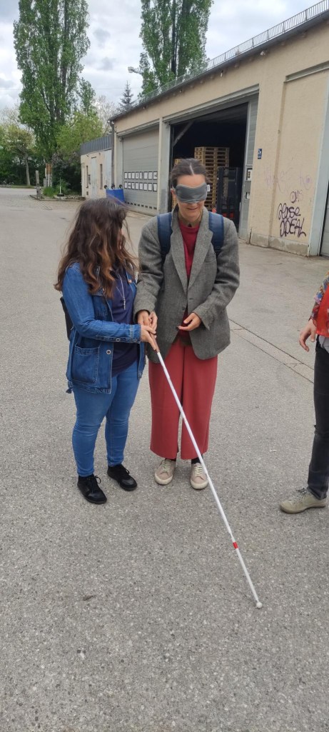 Eine blinde Person erklärt einer Person mit Schlafmaske wie ein Taststock funktioniert