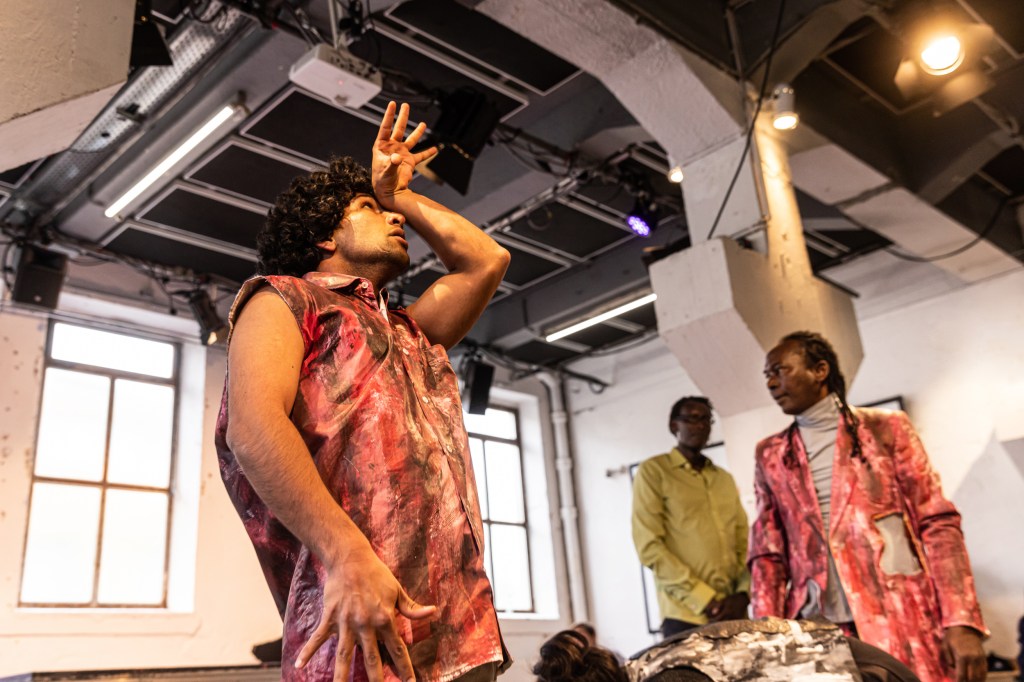 Eine Person of Colour in rot-brauner Weste hebt eine Hand an den Kopf, im Hintergrund eine weitere Person of Colour in rot-braunem Jackett und eine Person of Colour in grünem Hemd