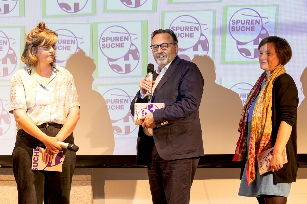 Zwei weiße Frauen und ein weißer Mann vor einer Leinwand, der Mann in der Mitte spricht in ein Mikrofon