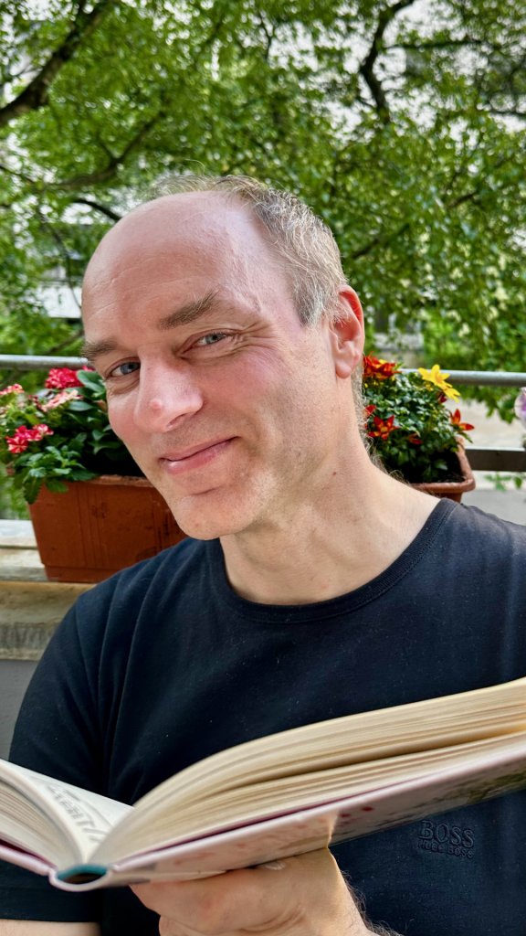 Moritz Staemmler hält ein Buch in der Hand und blickt lächelnd in die Kamera. Er trägt ein schwarzes T-Shirt. Im Hintergrund sieht man einen Pflanzenkübel und bunte Blumen.